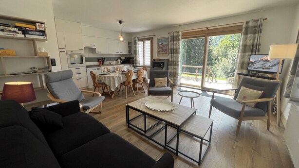 Una sala moderna con un sofá, una mesa de centro y un comedor con sillas y una mesa. Se observa una cocina al fondo con armarios y un microondas.