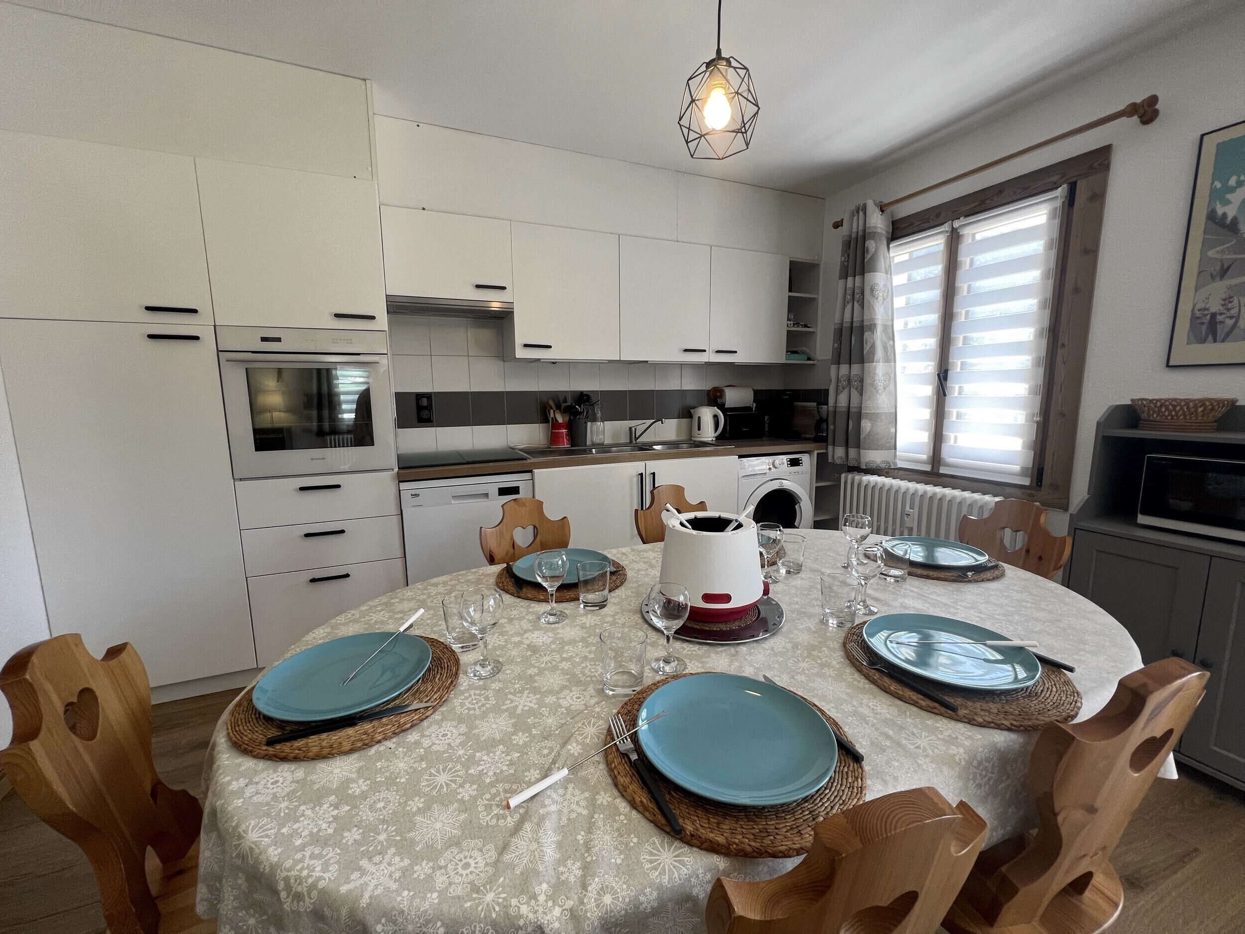 A modern kitchen with white cabinets, a dining table set for four, and a window with blinds.