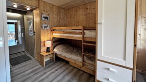 Een slaapkamer met een houten stapelbed, een klein nachtkastje met lamp, een witte kast en een houten deur die naar een andere kamer leidt.