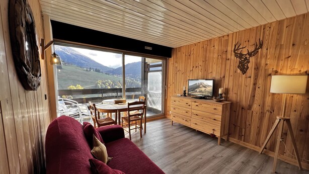 Een knusse woonkamer met een rode bank, houten wanden, een televisie op een houten kast en een prachtig uitzicht door grote ramen.