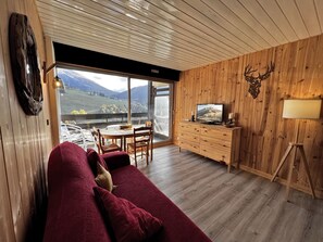 A cozy living room with a red sofa, wooden walls, a TV on a wooden cabinet, and a scenic view through large windows. - AIGUILLE VERTE 28 - Studio with mountain views, close to the aquatic area (La Clusaz)