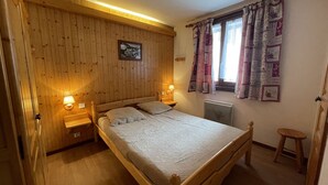 Une chambre à coucher aux murs lambrissés, avec un lit, deux lampes de chevet, une fenêtre ornée de rideaux à motifs et un radiateur.