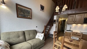 A cozy living room with a sofa, a dining table with chairs, a staircase, and a kitchen area. - RICHEMONT 12 – Duplex near the slopes with village views – Garage (La Clusaz)