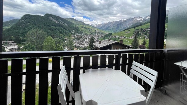 A balcony with a white table and chairs, overlooking a mountainous landscape with a village below. - RICHEMONT 12 – Duplex near the slopes with village views – Garage (La Clusaz)