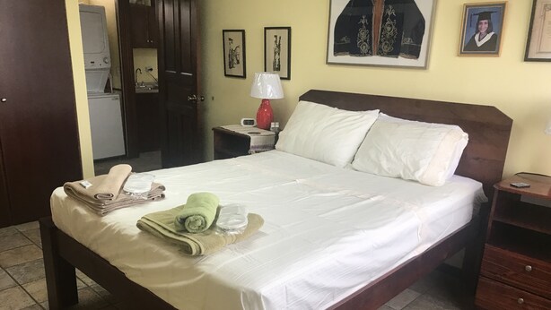 A neatly made bed with white linens, a green towel, and a white pillow. There is a wooden nightstand with a red lamp and a clock. - Deluxe Two-Bedroom Penthouse Apartment in Escalante, San Jose Center (San José)