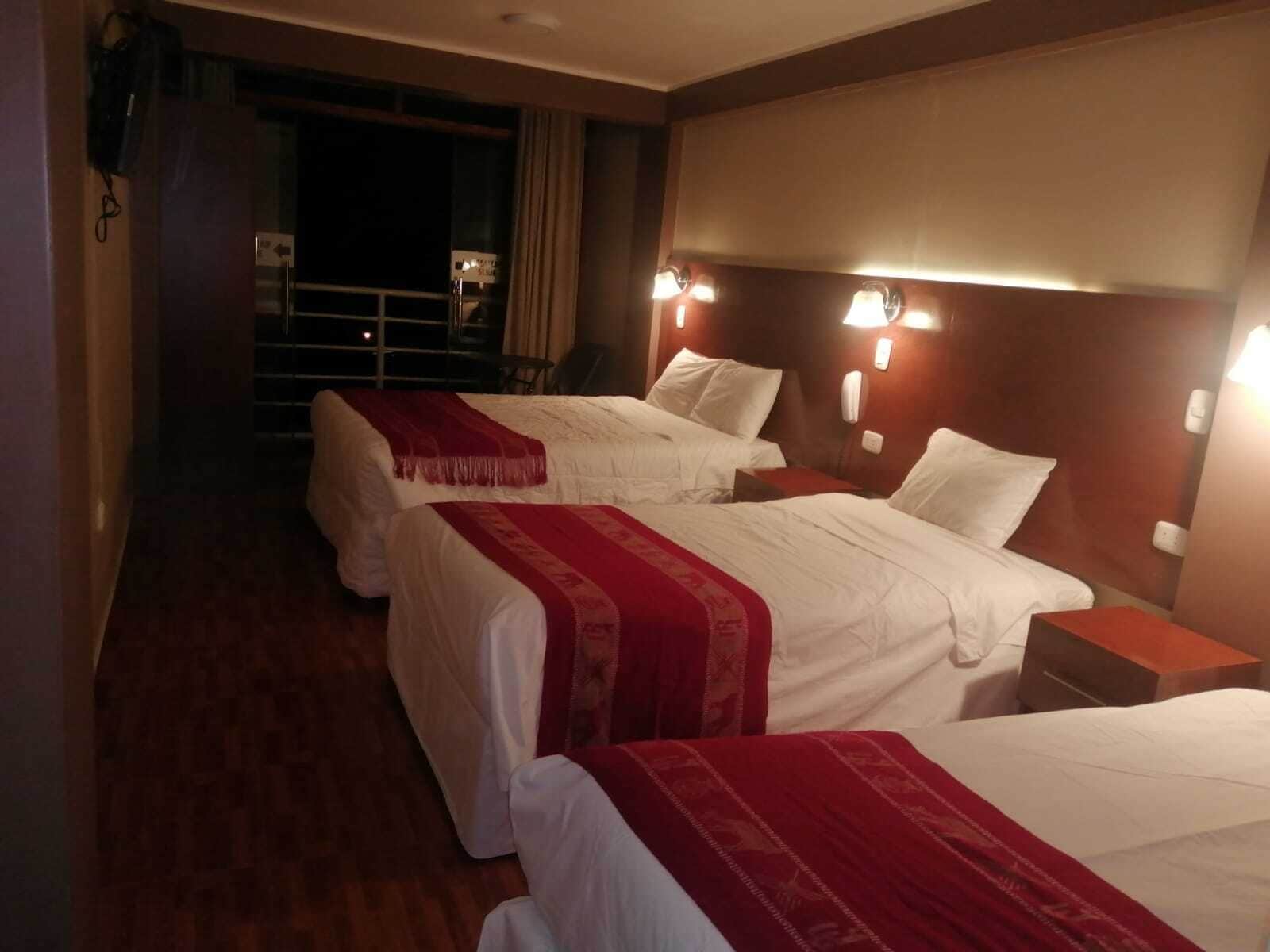 A hotel room with three beds, each with white bedding and red throw blankets. There are bedside lamps and a nightstand with a lamp. The room has wooden flooring and a dark-colored wall.