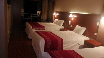 A hotel room with three beds, each with white bedding and red throw blankets. There are bedside lamps and a nightstand with a lamp. The room has wooden flooring and a dark-colored wall.