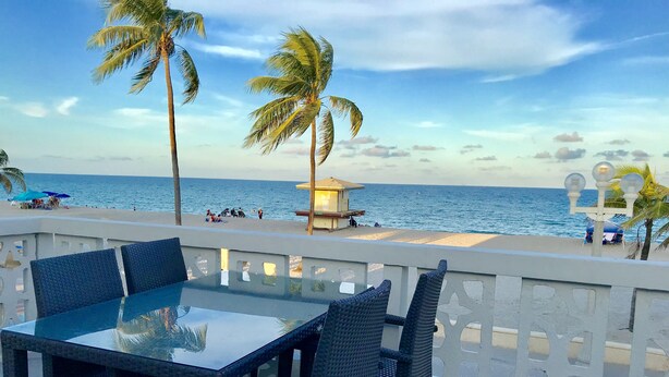 Un tavolo con piano in vetro e sedie in vimini su un balcone con vista su una spiaggia, palme e una torretta di salvataggio.