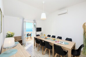 Een eetruimte met een houten tafel, zwarte stoelen, een witte hanglamp en uitzicht op zee door een open deur.