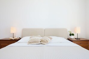 Een net opgemaakt bed met een witte dekbedovertrek en twee decoratieve kussens, met aan weerszijden nachtkastjes met lampen en een kleine plant.