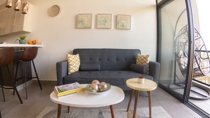 Un salon moderne avec un canapé gris, deux tables blanches et un coin cuisine doté de meubles en bois et d’un tabouret de bar.