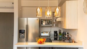 Une cuisine moderne équipée d’un réfrigérateur en acier inoxydable, d’un four à micro-ondes, d’un grille-pain et d’un évier avec un robinet en cuivre. Des lampes suspendues et un saladier rempli de fruits ornent le plan de travail.