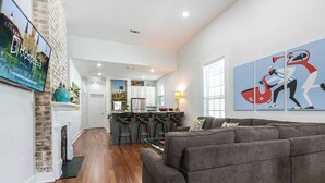A modern living room with a grey sofa, a fireplace, and a kitchen in the background. - 15% OFF Monthly | 10% OFF Weekly-Spacious 5BR Near Magazine St & Uptown (New Orleans)
