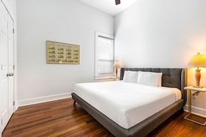 Une chambre à coucher avec un grand lit, une table de chevet, une lampe, un tableau encadré au mur et une fenêtre avec des stores.