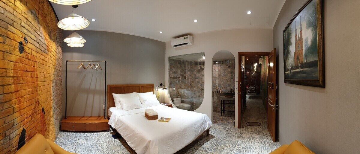 Une chambre d’hôtel avec un mur en briques, un lit recouvert de draps blancs, un miroir rond et un tableau accroché au mur.