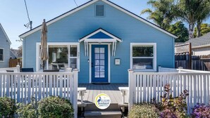 A blue house with a front porch, white picket fence, and a logo for Beachnest Vacation Rentals. - 8th Avenue Beach Cottage (Santa Cruz)