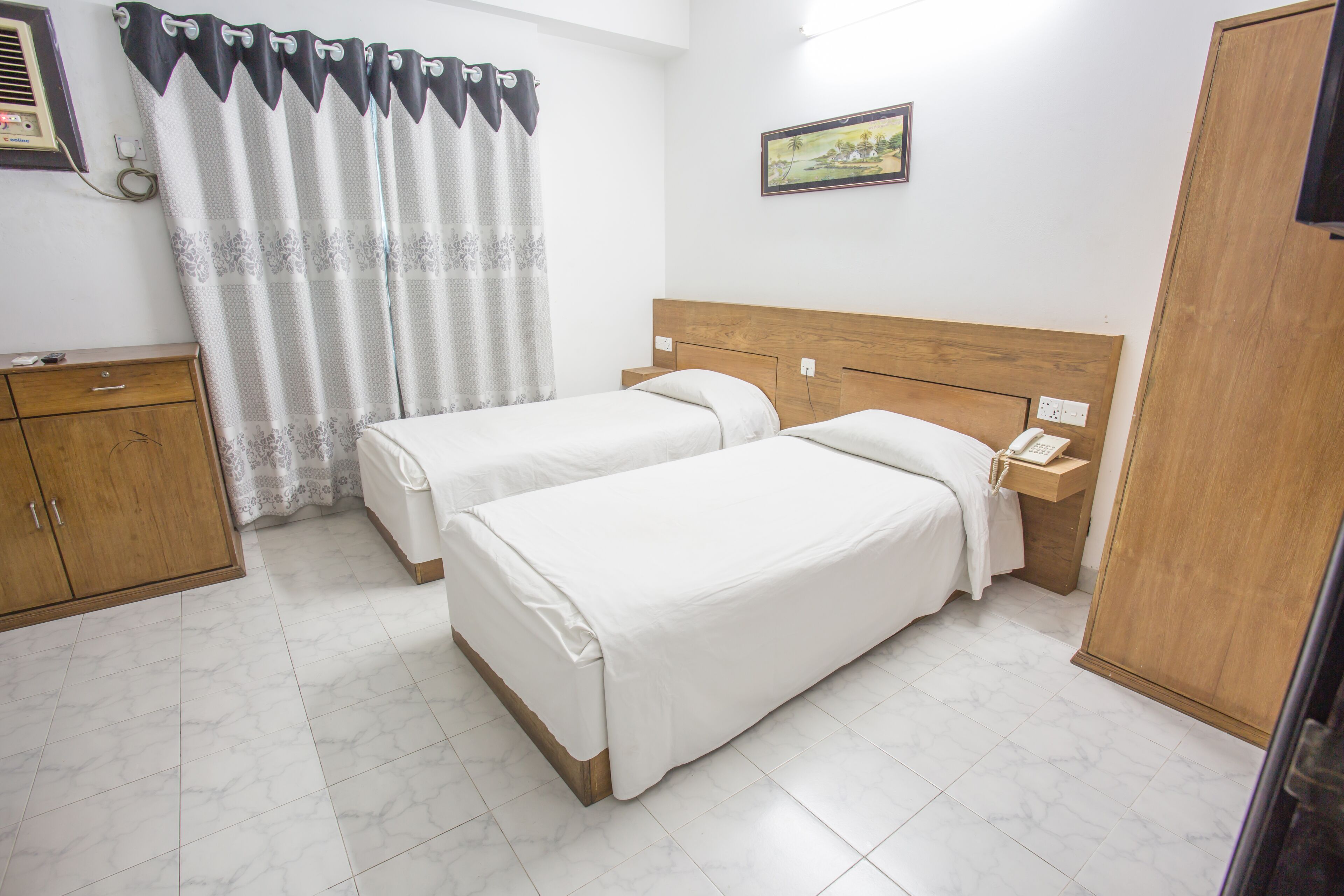 A hotel room with two beds, a wooden headboard, a nightstand with a telephone, a wooden wardrobe, and a window with a curtain.