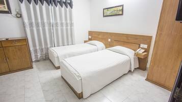 A hotel room with two beds, a wooden headboard, a nightstand with a telephone, a wooden wardrobe, and a window with a curtain.