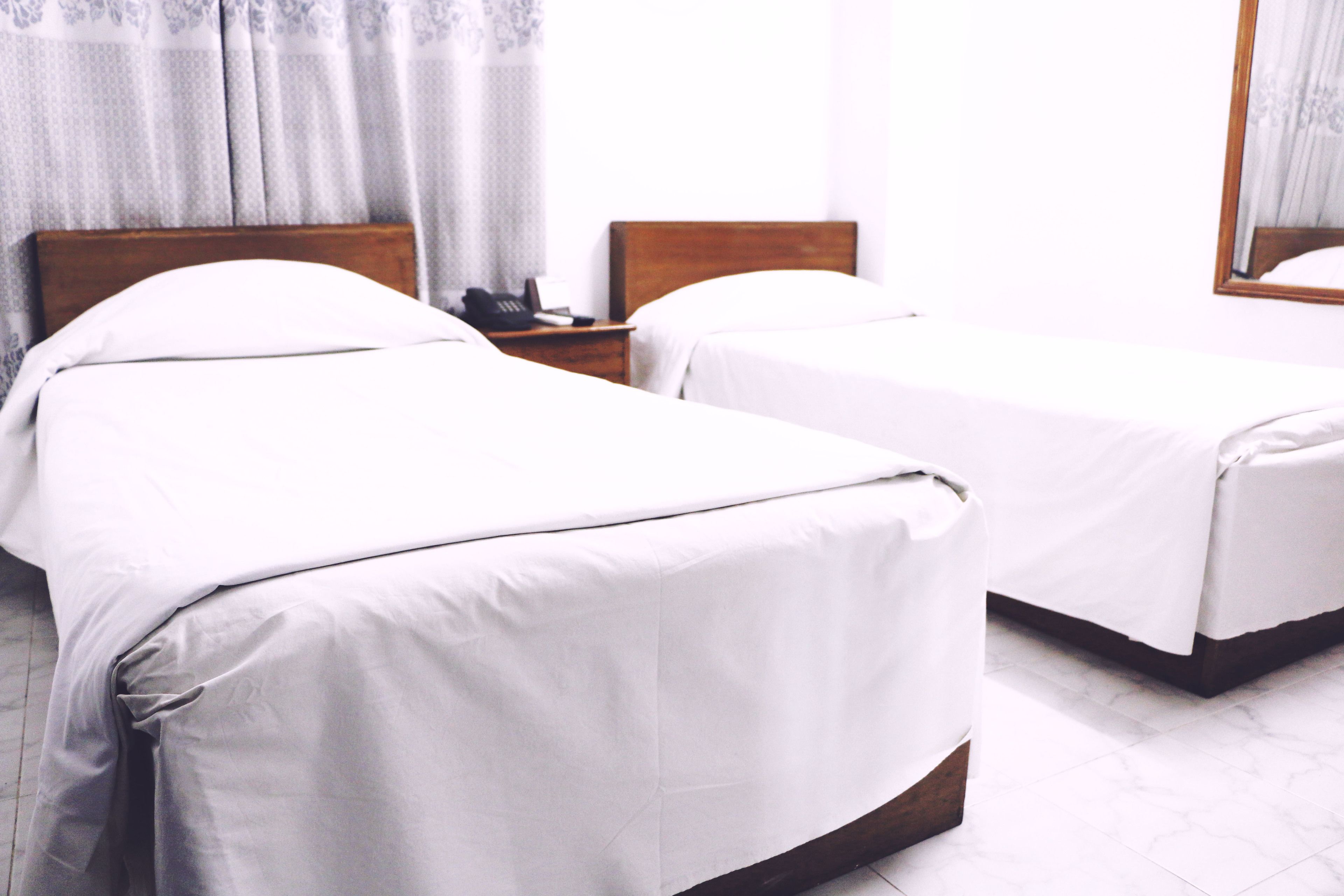 A hotel room with two single beds, a wooden headboard, a bedside table with a phone, and a large mirror on the wall.