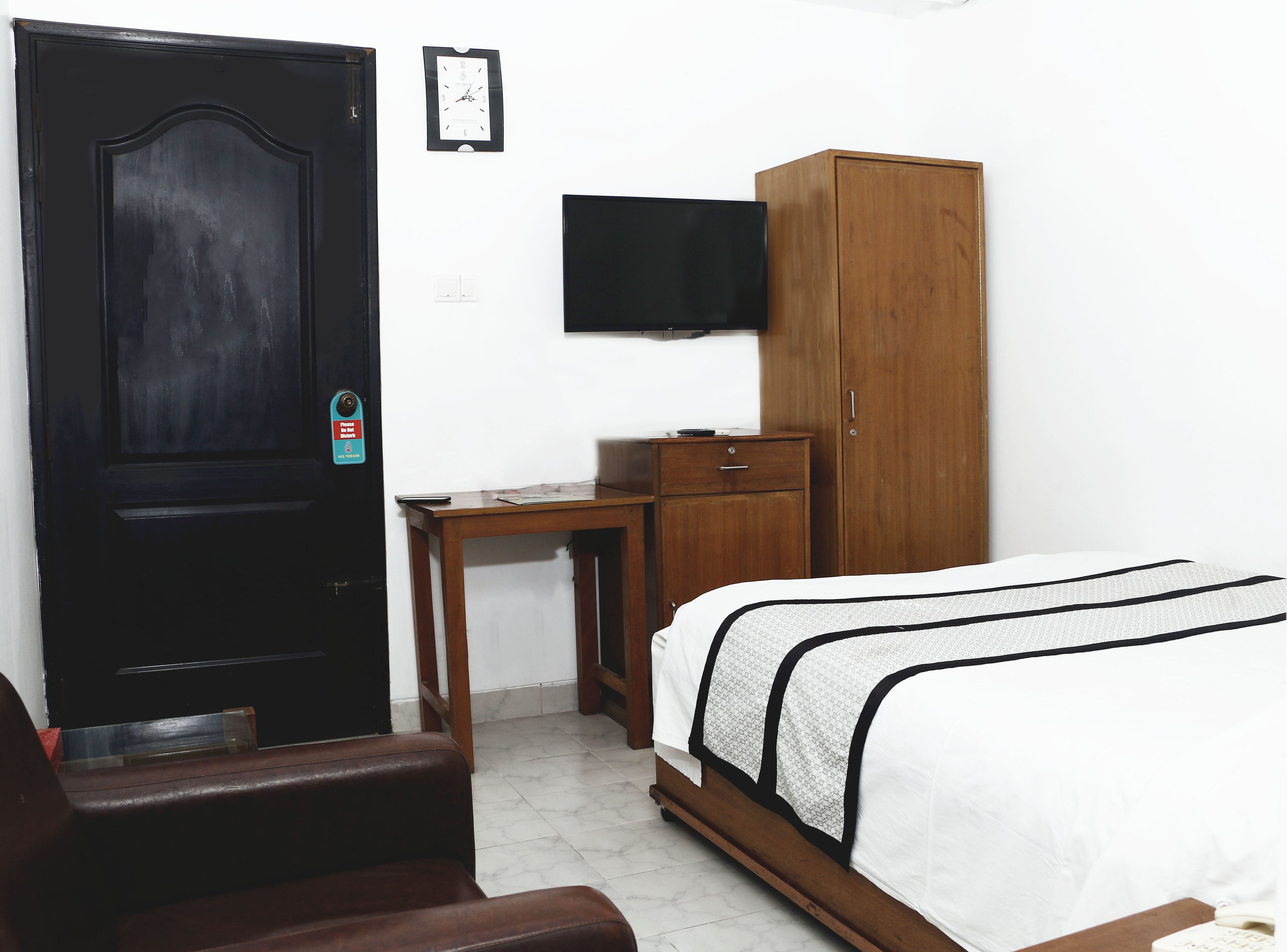 A bedroom with a bed, a wooden wardrobe, a desk, a chair, and a television mounted on the wall.