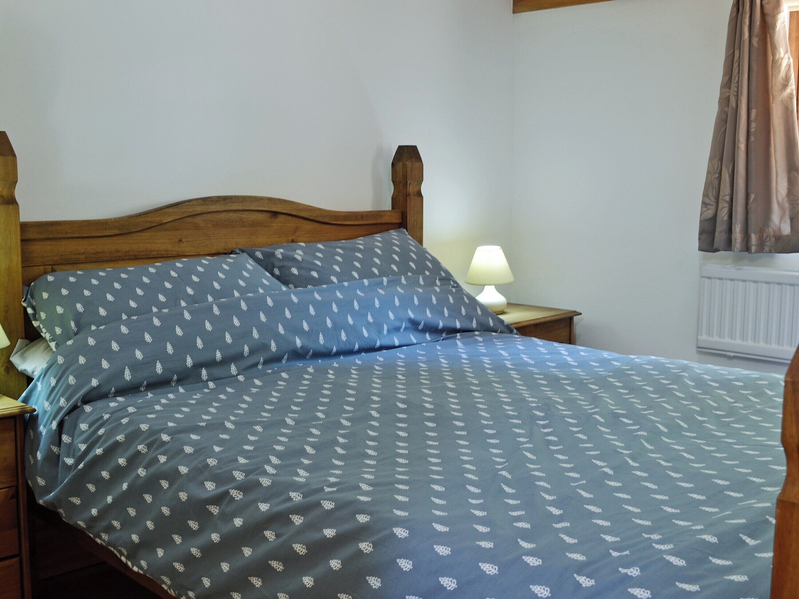 A bed with a patterned blue and white bedspread, a wooden headboard, a bedside lamp, and a window with curtains.