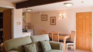 A cozy living room with a green sofa, wooden dining table, and chairs. There is a kitchen area with cabinets and a kettle. - 1 bedroom accommodation in Sidbury, near Sidmouth (Sidbury, near Sidmouth)