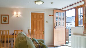A cozy living room with a green sofa, wooden chairs, a dining table, and a large window with a view of buildings outside. - 1 bedroom accommodation in Sidbury, near Sidmouth (Sidbury, near Sidmouth)