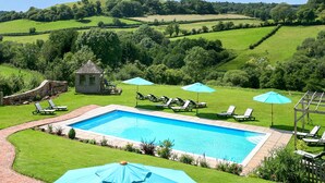 A rectangular swimming pool with a brick border, surrounded by green grass and a few trees. There are lounge chairs and umbrellas around the pool. - 1 bedroom accommodation in Sidbury, near Sidmouth (Sidbury, near Sidmouth)