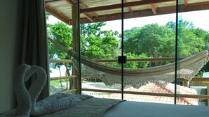 A bed with a white pillow and a textured blanket, a hammock outside a window, and a view of trees and a thatched roof structure. - Pousada Maré (Prado)