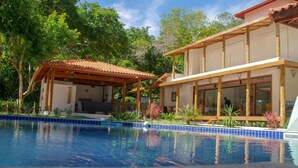 A poolside area with a wooden deck, a covered seating area, and a building with large windows. - Pousada Maré (Prado)