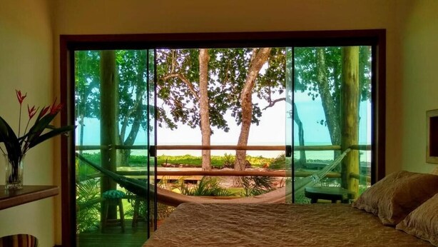 A bedroom with a large bed, a desk with a chair, and a view of a hammock and trees through large glass doors. - Pousada Maré (Prado)