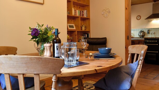 A dining area with a wooden table, chairs, a vase with flowers, a wine bottle, and a bookshelf in the background. - 1 bedroom accommodation in Lindale, near Grange-over-Sands (Lindale, near Grange-over-Sands)