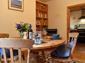 A dining area with a wooden table, chairs, a vase with flowers, a wine bottle, and a bookshelf in the background.