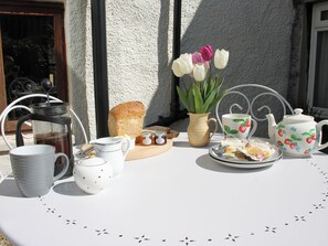 Una mesa dispuesta con una cafetera, tetera, tazas, un plato con pasteles, un florero con flores y un pan.