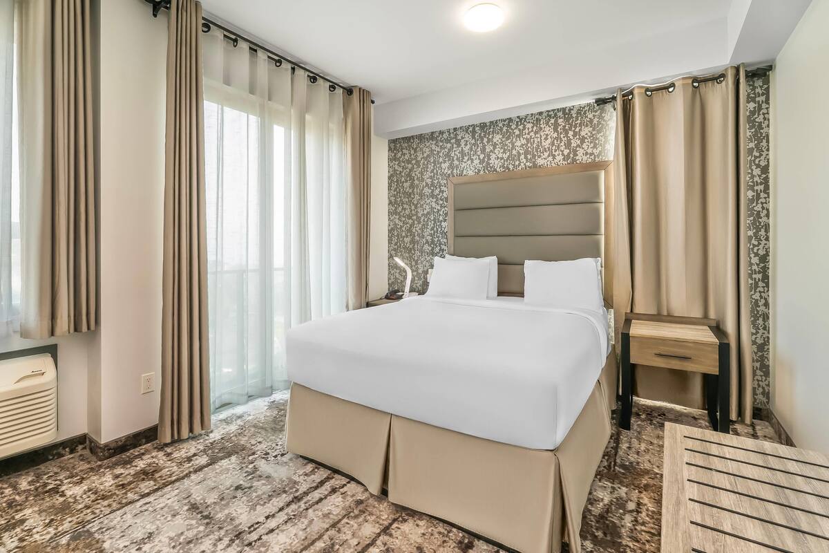 A hotel room with a large bed, a bedside table, a nightstand, a window with curtains, and a patterned wall.