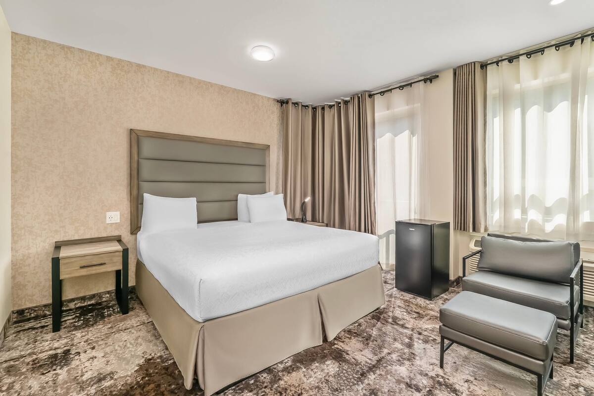 A hotel room with a large bed, a bedside table, a chair, and a small black speaker.
