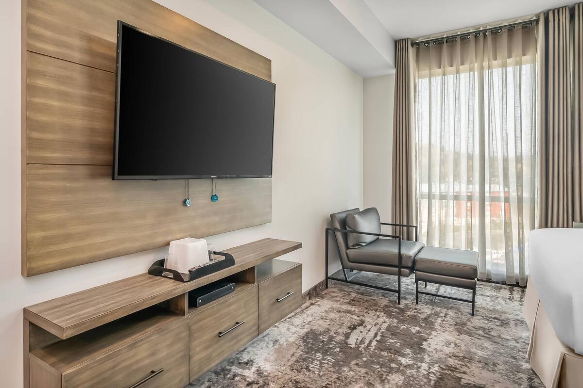 A hotel room with a flat-screen TV mounted on a wooden panel wall, a seating area with a chair and ottoman, a large window with curtains, and a bed with a white cover.