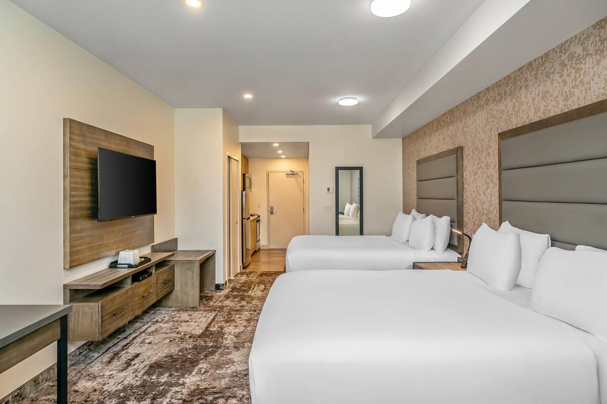 A hotel room with two beds, a TV mounted on the wall, a wooden console table, and a patterned carpet.