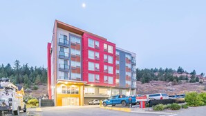 Un hotel de varios pisos con fachada roja y gris, una entrada a nivel de calle y un estacionamiento con varios vehículos.