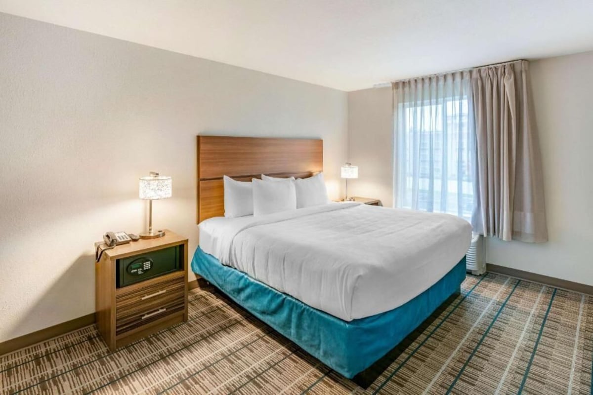 A hotel room with a bed, bedside lamps, a nightstand with a phone and a small drawer, and a window with curtains.