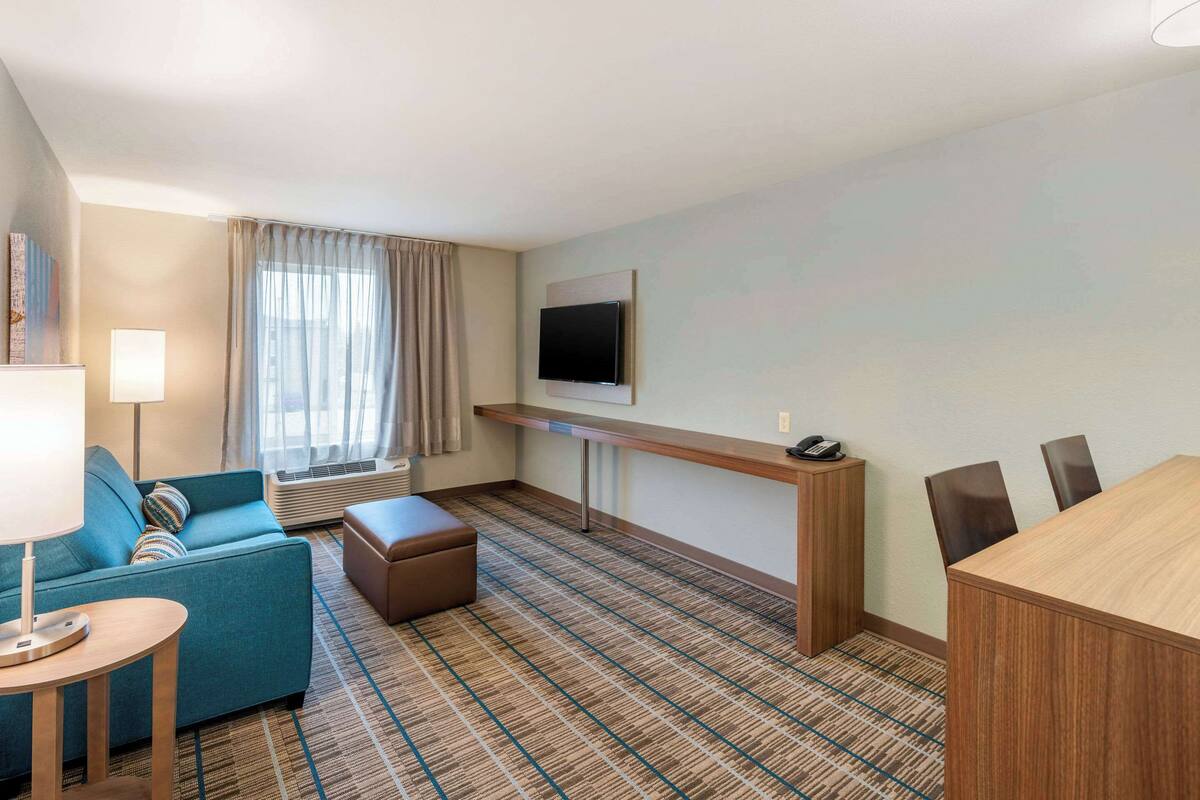 A hotel room with a sofa, a small table, a TV mounted on the wall, and a carpeted floor.