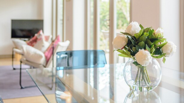 Een moderne woonkamer met een glazen salontafel, een transparante vaas met witte bloemen en een bank met kussens.
