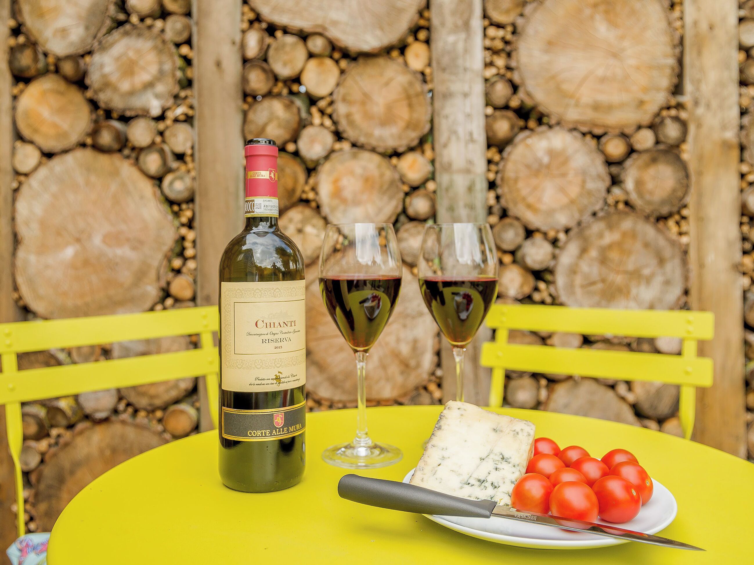 Une bouteille de vin, deux verres, une assiette de fromage et des tomates sur une table jaune, avec un mur de pierres en arrière-plan.