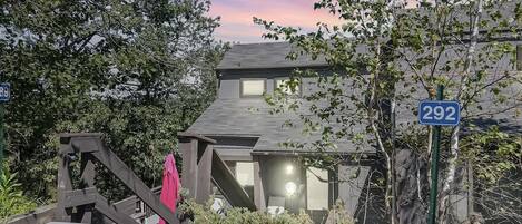 Una casa de dos pisos con el número '292' visible en un cartel, rodeada de follaje y una escalera de madera.