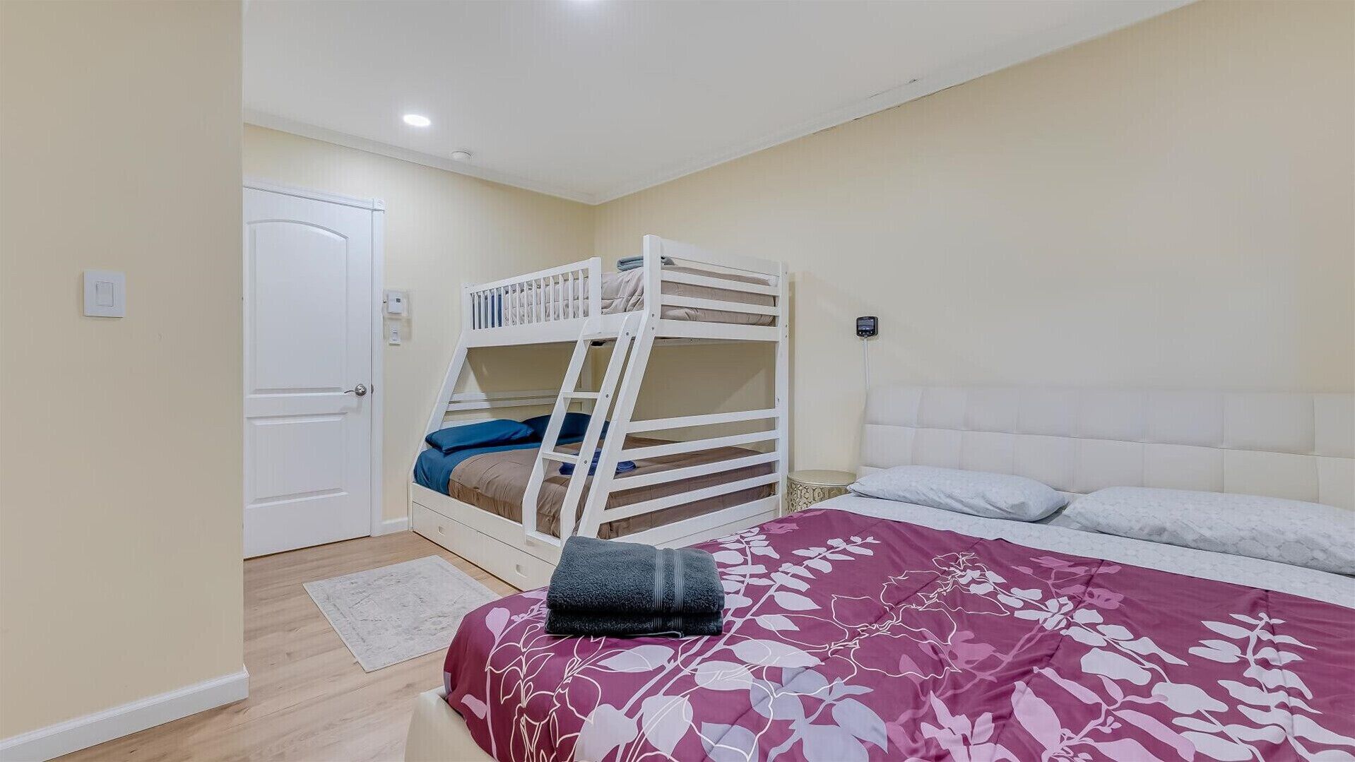 A bedroom with a bunk bed, a single bed, a white door, and a bedside table with a lamp.