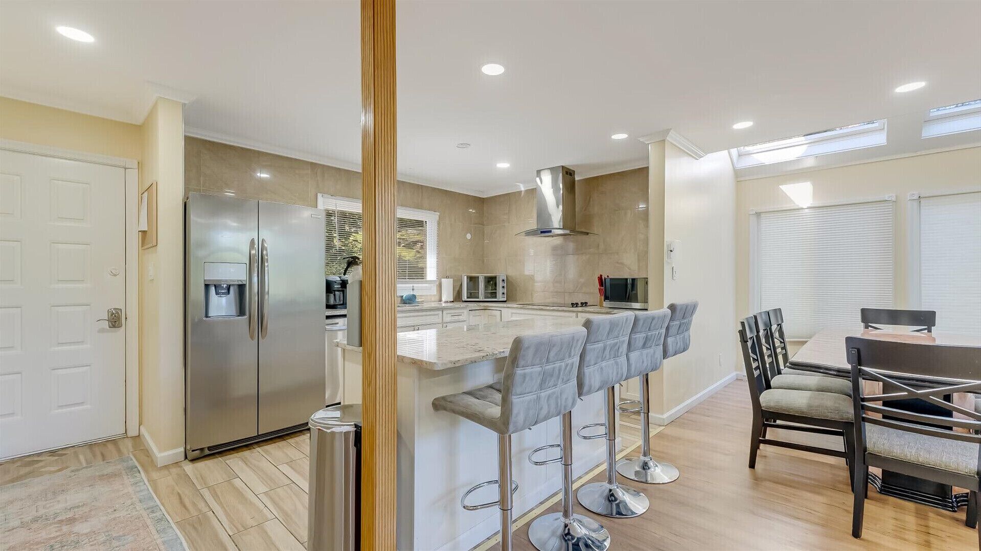 Una cocina moderna con un refrigerador de acero inoxidable, un horno empotrado y un microondas. Hay taburetes para bar y una mesa de comedor con sillas.