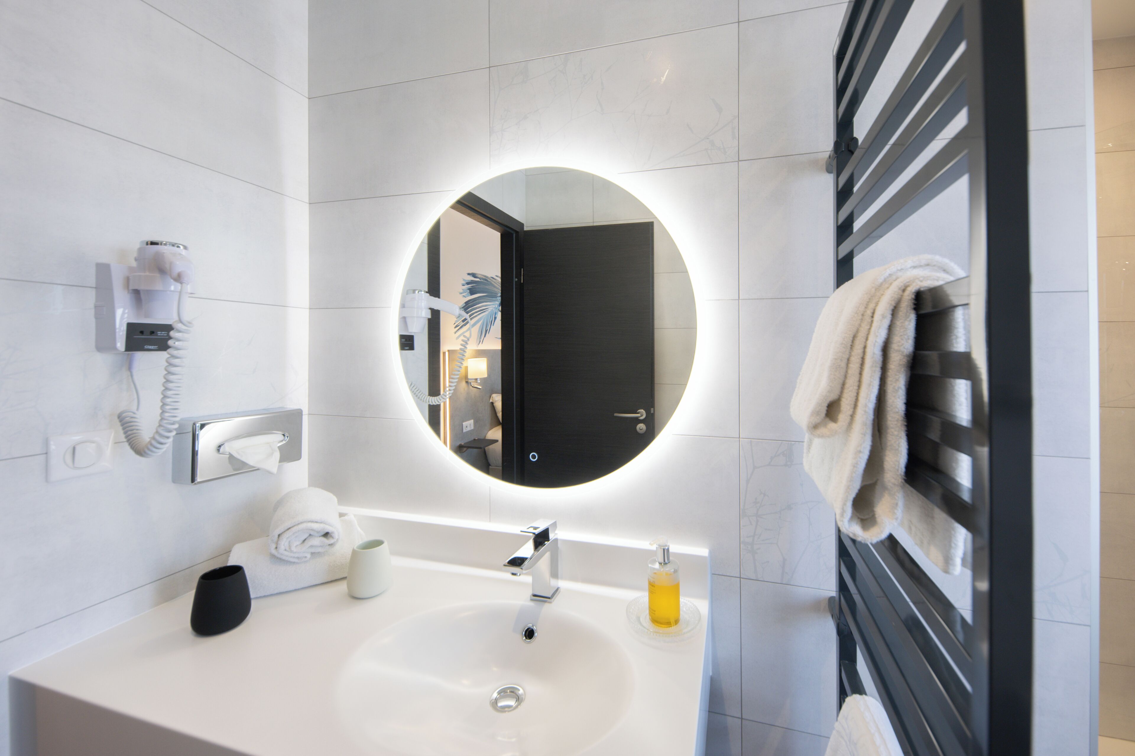 A modern bathroom with a round mirror, a sink, a towel rack, a telephone, and a soap dispenser.