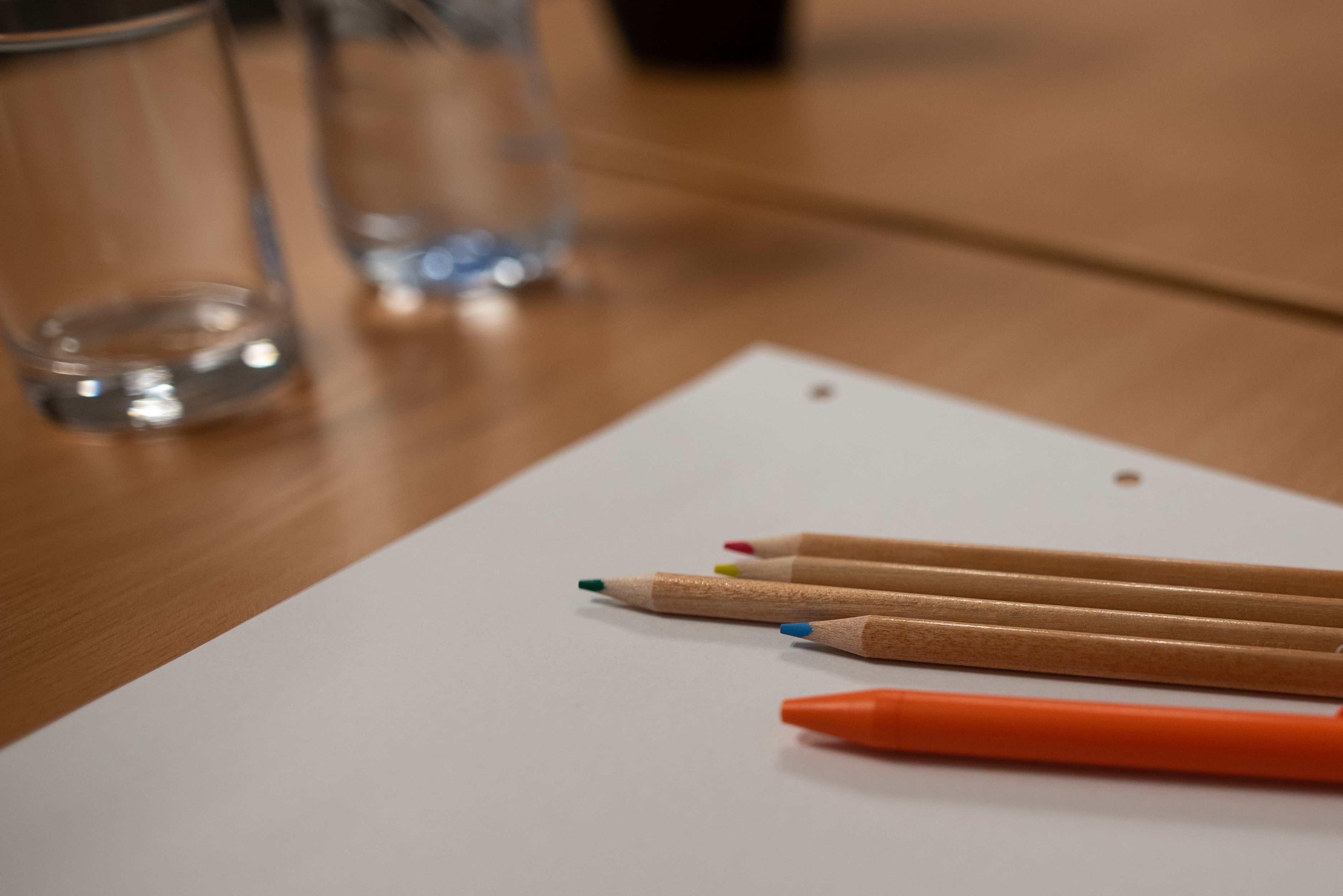 Ein Holztisch mit einem weißen Papierbogen, mehreren farbigen Stiften und einem orangefarbenen Stift.