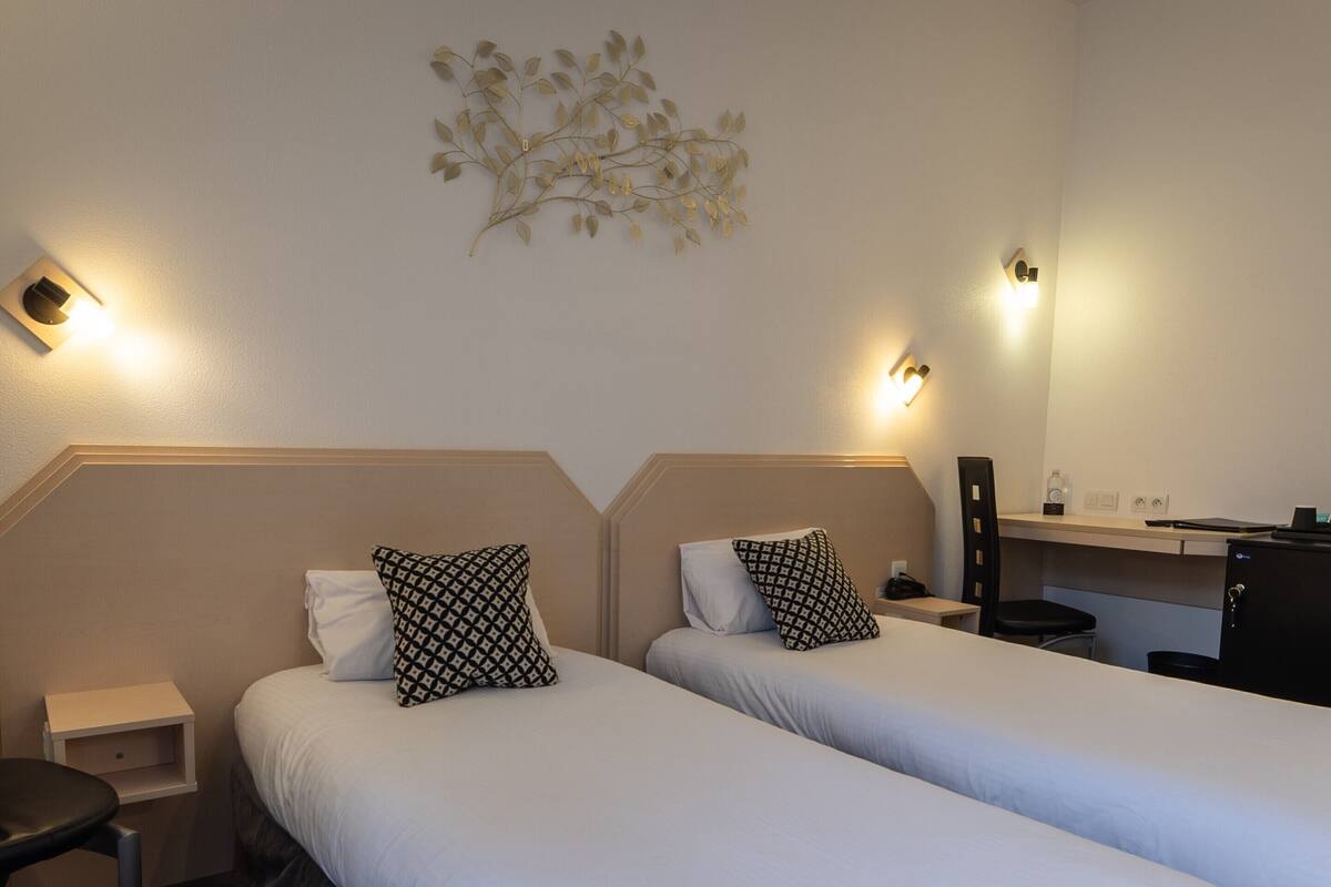 Ein Hotelzimmer mit zwei Betten, einem kleinen Schreibtisch, einem Stuhl, einem Nachttisch und Wandleuchten.