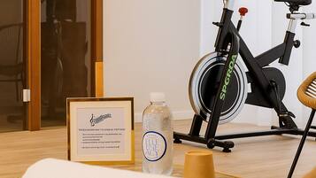 Una habitación con una silla de mimbre, una toalla, un certificado enmarcado, una botella y una bicicleta estática.